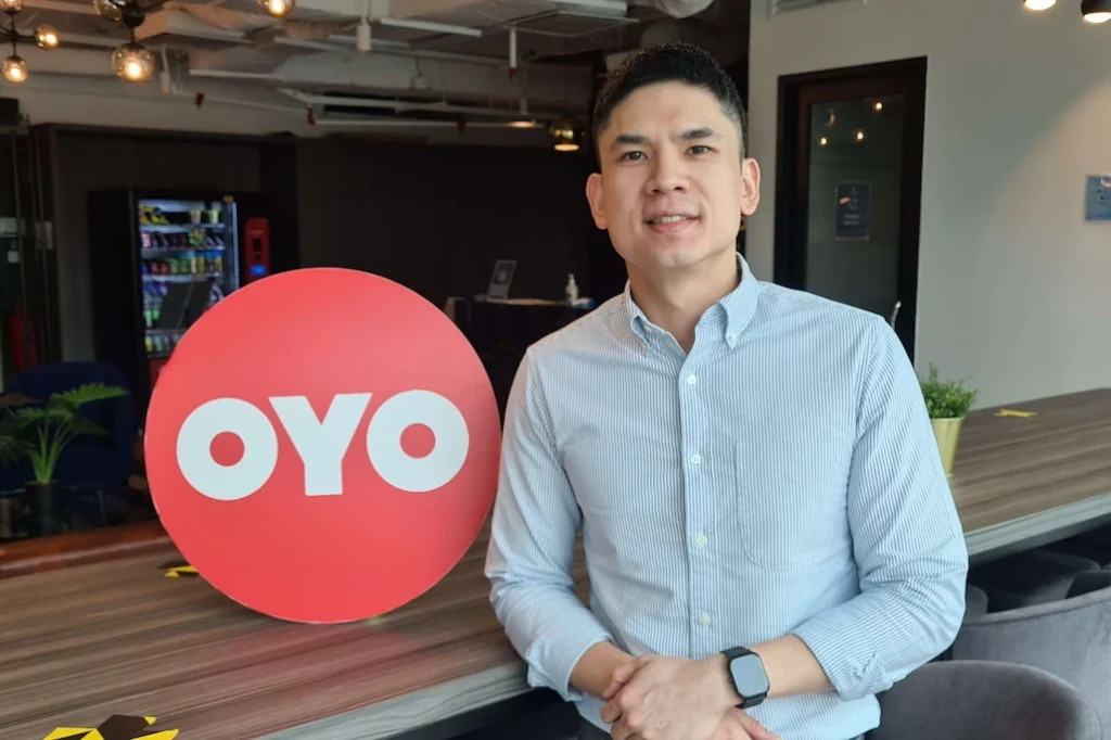 OYO Hotels regional lead standing beside the red OYO logo sign in a modern office
