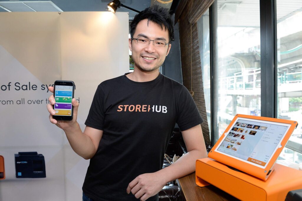 StoreHub founder holding a smartphone beside the company's orange point-of-sale terminal