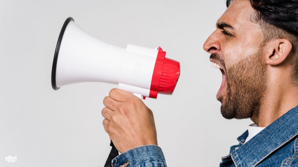 Man in a denim jacket shouting into a red and white megaphone against a plain background