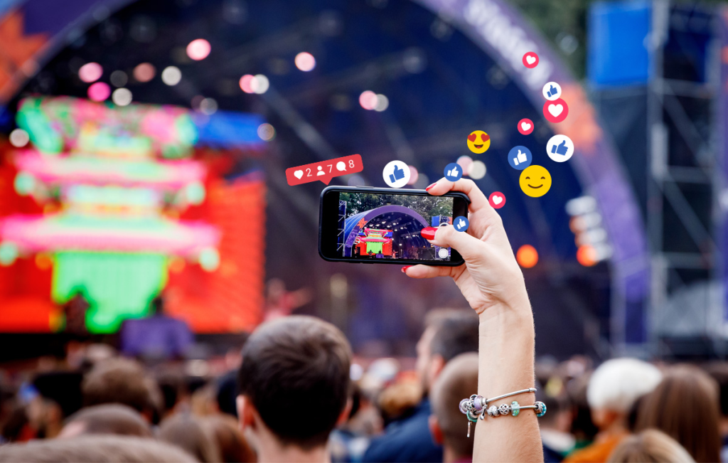 Hand holding a phone filming a concert stage with social media like and heart icons floating above the crowd