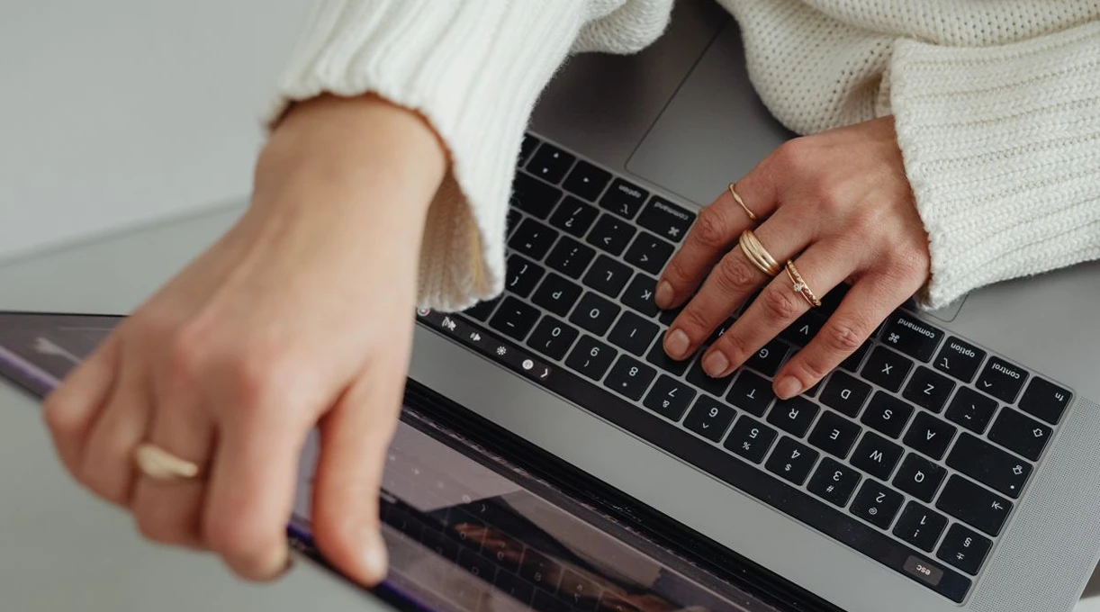 Hands wearing gold rings typing on a laptop keyboard on a white desk