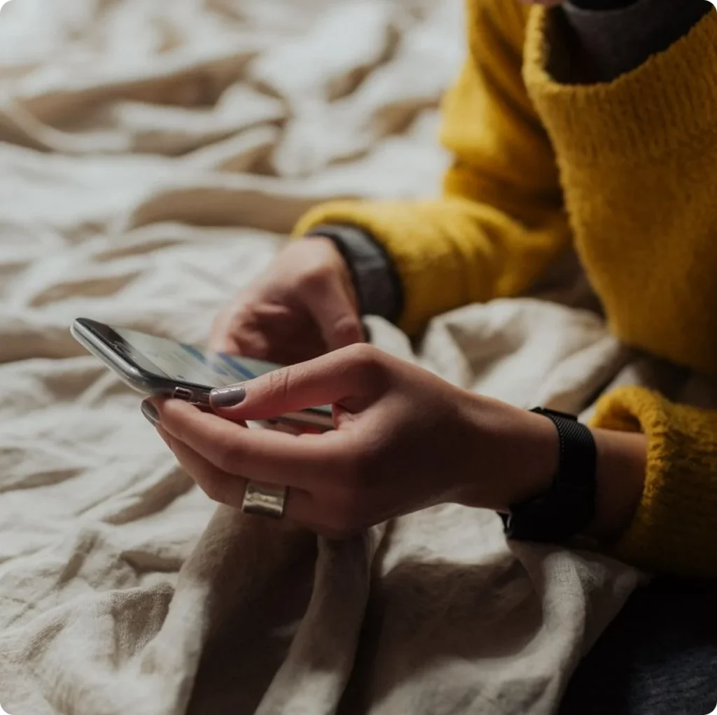 Woman in a yellow sweater scrolling social media on a smartphone while relaxing on a bed