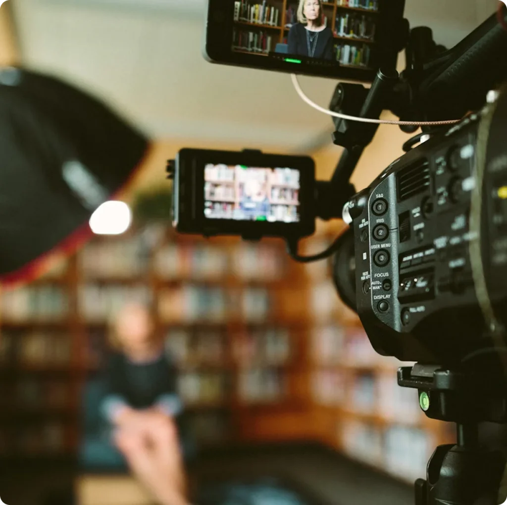 Video camera and monitor framing a woman during a media interview inside a library setting