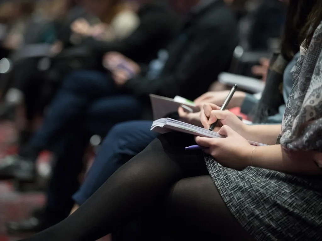 Audience members taking notes in notebooks during a press conference or industry event