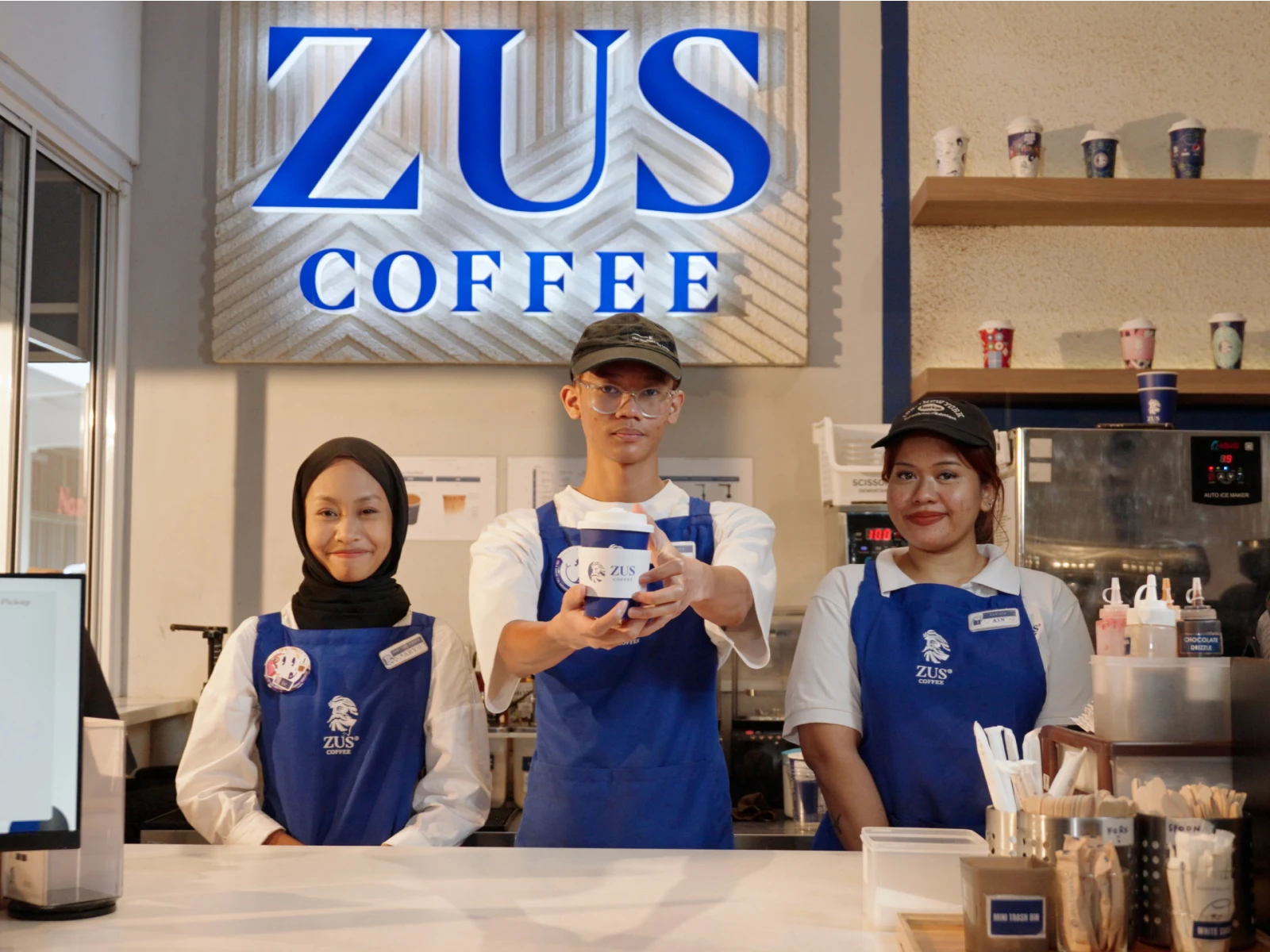 Zus Coffee baristas in blue aprons serving a drink at the counter under the brand sign